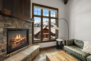 Ein Kamin mit Steinverkleidung, ein Fenster mit Blick auf einen verschneiten Berg, ein grünes Ledersofa und ein Holztisch.