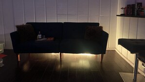 En mørk sofa med to puder, et lille bord med en flaske og glas, samt en reol med bøger og pynteting.