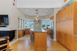 A modern kitchen with wooden cabinets, a central island, a built-in oven, and a large window providing natural light. - 2nd Row Designer Cottage w/ Ocean Views, Elevator & Hot Tub (Emerald Isle)
