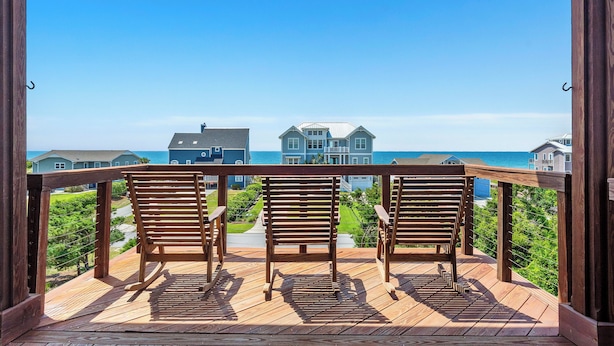 Een houten terras met drie stoelen, gericht op een groot huis met een blauw dak en uitzicht op de oceaan.
