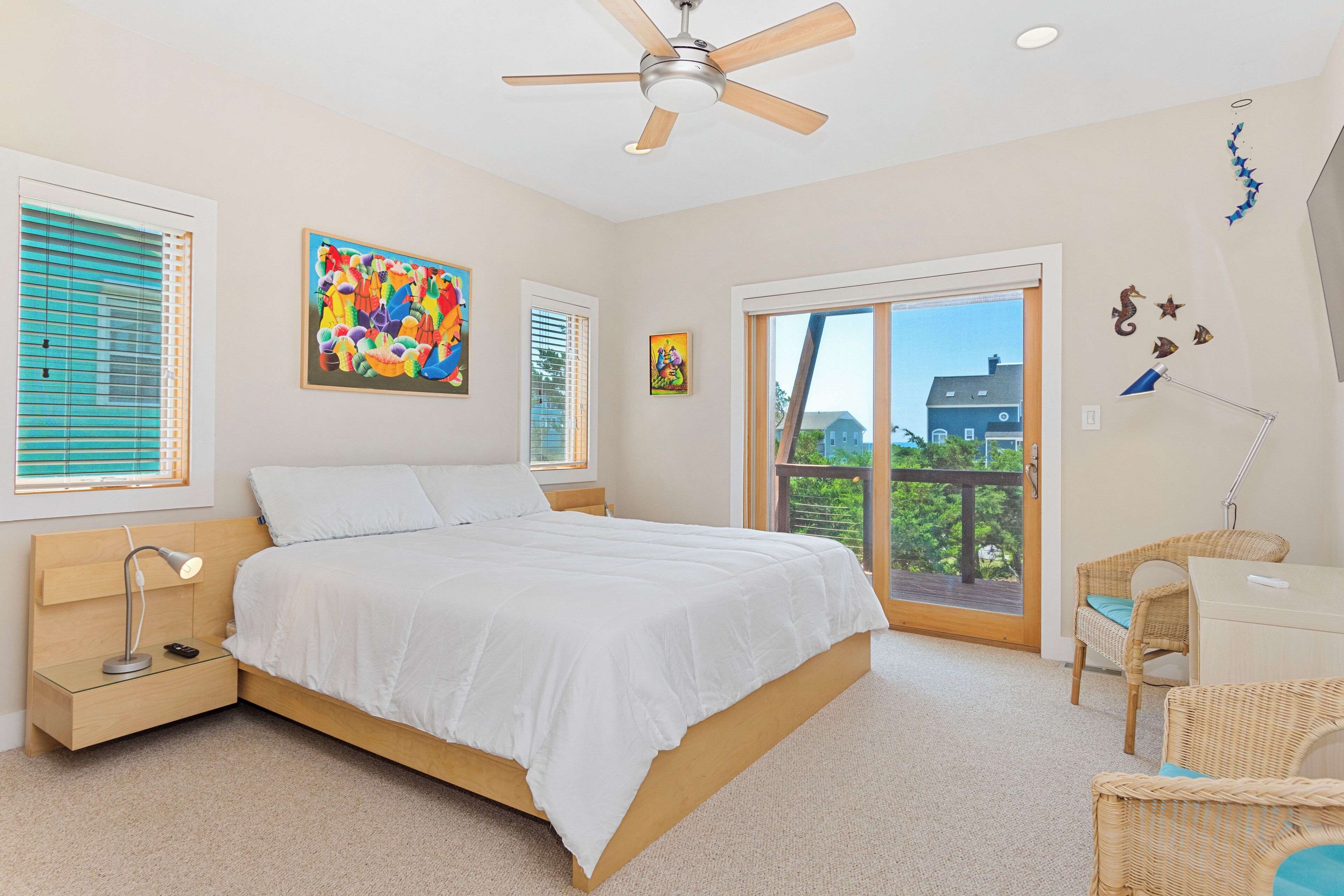 A bedroom with a bed, a ceiling fan, a desk, wicker chairs, and a balcony with a view.