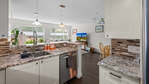 A modern kitchen with granite countertops, stainless steel appliances, pendant lighting, and a view of greenery outside.