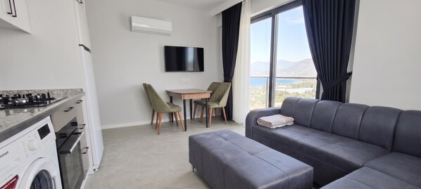 A modern living room with a grey sofa, a small table, and chairs. A washing machine is visible in the corner. - Doğuş Apart (Antalya)