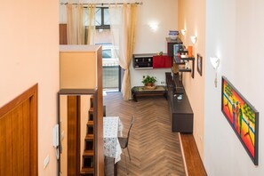 Ein kompakter Wohnbereich mit einer Holztreppe, einer Essecke mit Tisch und Stühlen, einer Küchenzeile und einem großen Fenster mit Vorhängen.