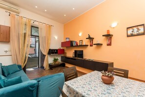Ein Wohnzimmer mit einer Couch, einem Esstisch mit gemustertem Tischlauf, einem Fernseher und einer Küchenzeile mit Schränken und einer Arbeitsplatte.