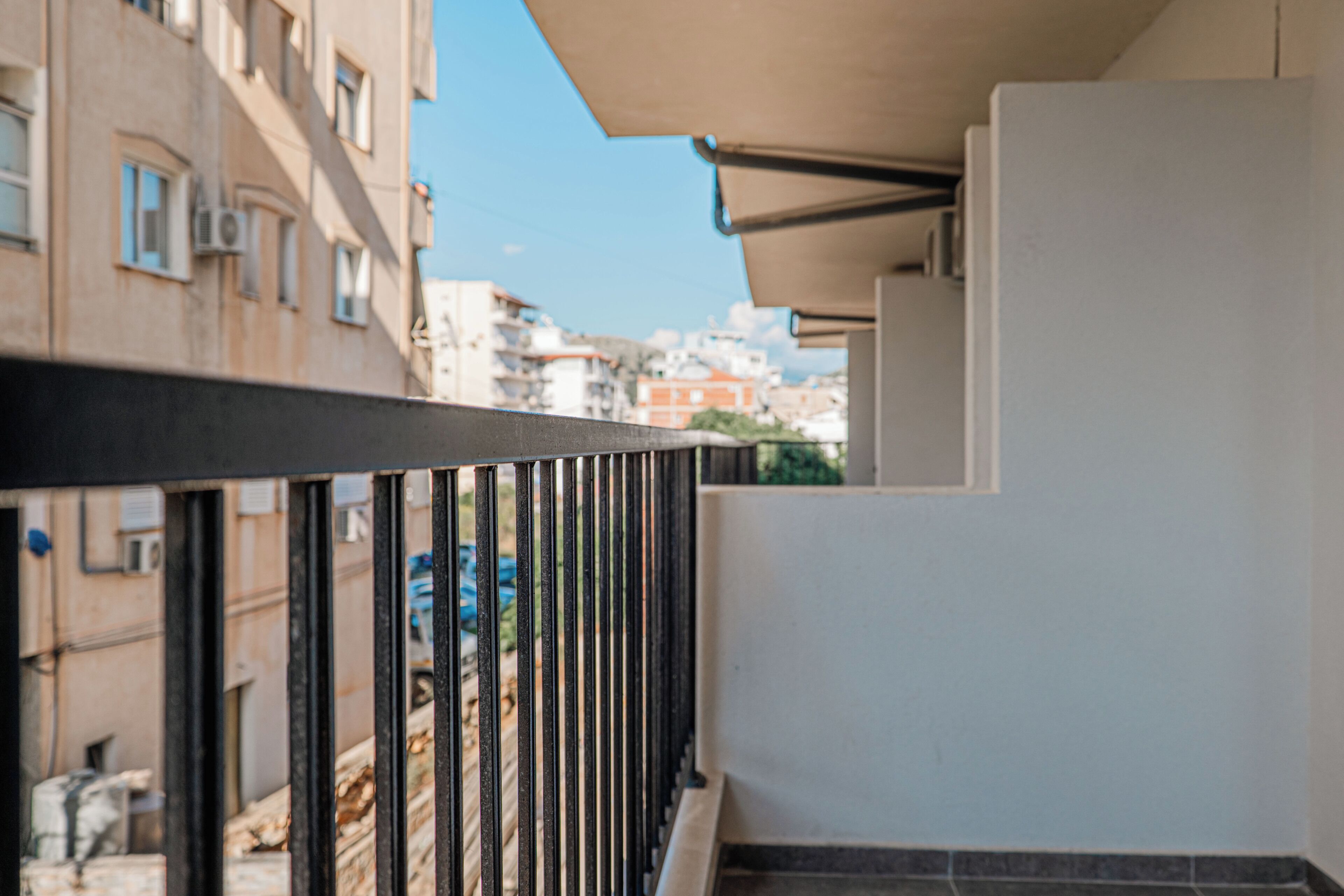 A balcony with a metal railing, a white wall, and a view of buildings in the background.