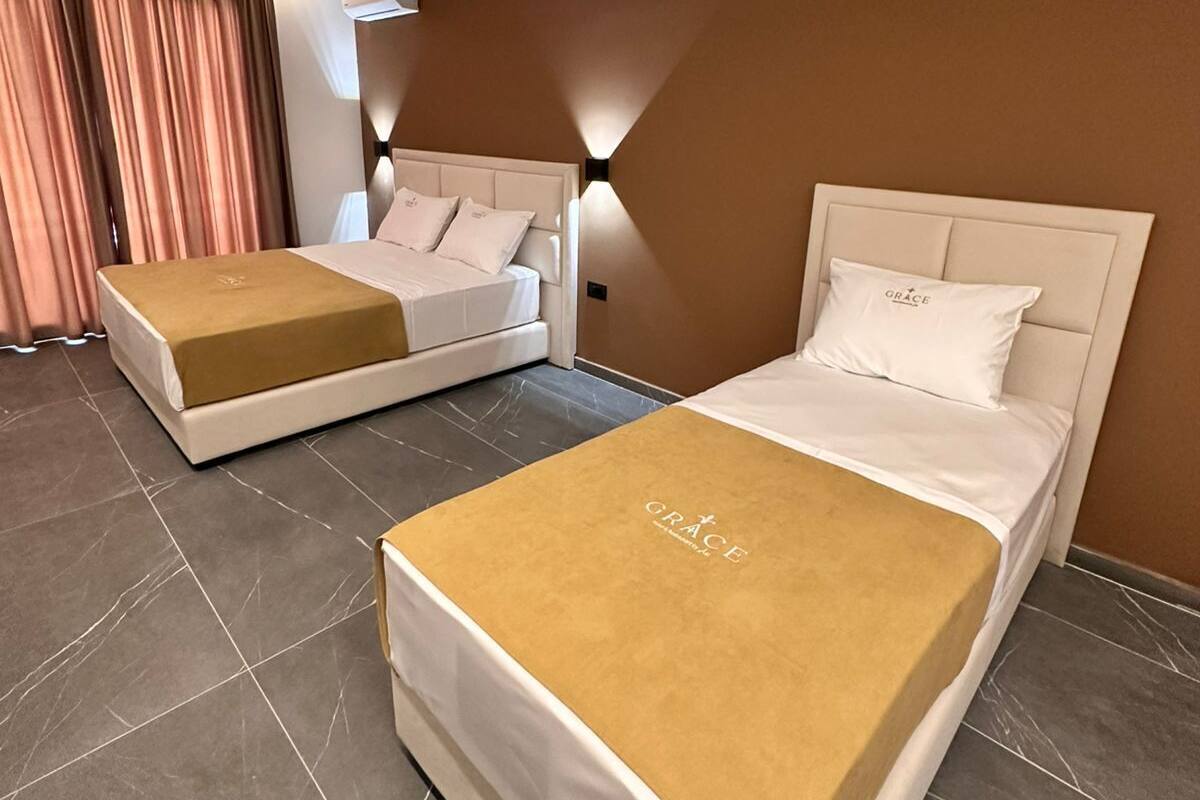 A hotel room with two beds, a large window with curtains, a wall-mounted air conditioner, and a light fixture.