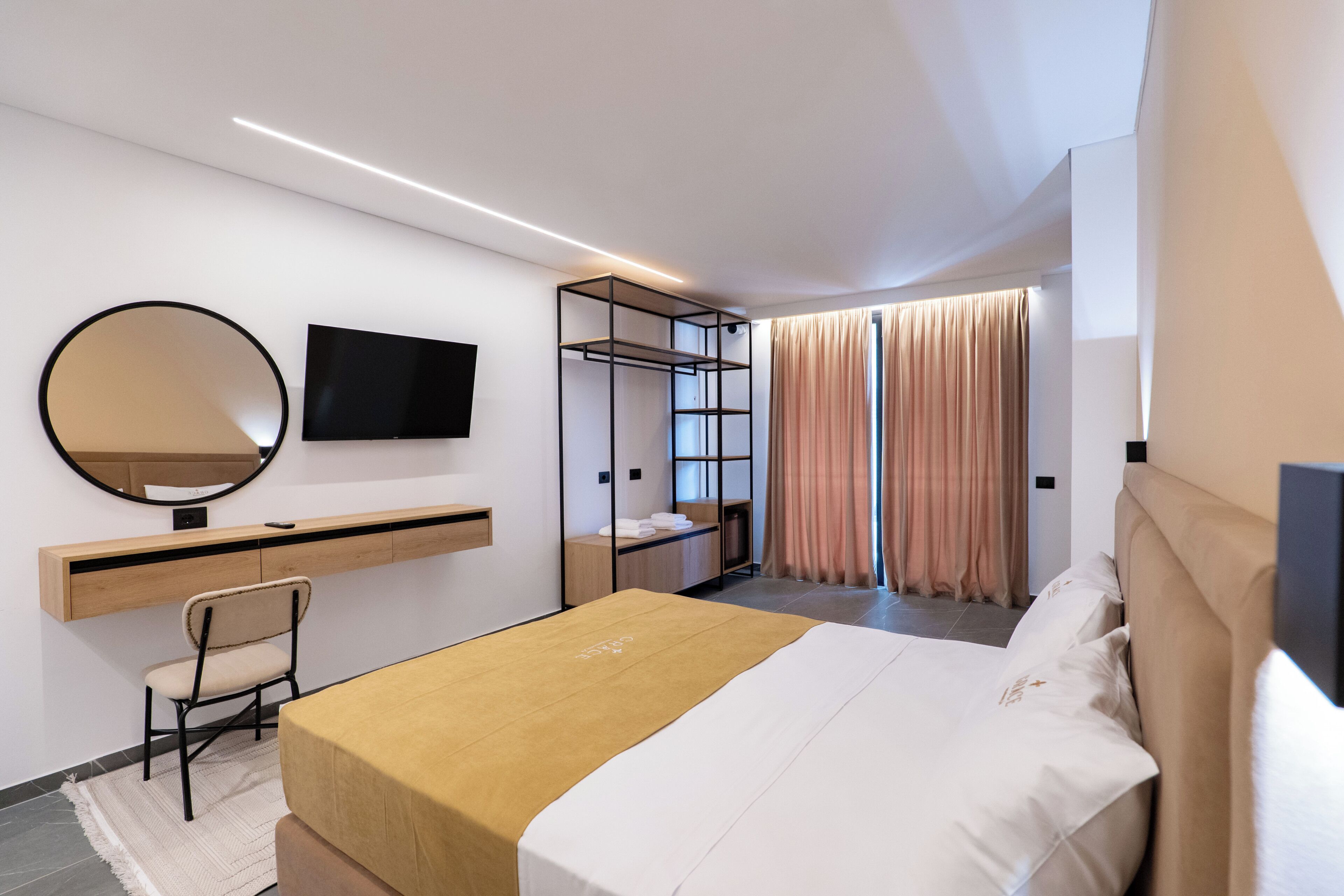 Una camera d'albergo moderna con un letto, una scrivania con una sedia, una TV, uno specchio grande e una finestra con tende.