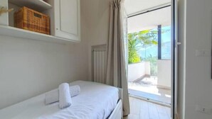 A single bed with a white mattress and a rolled towel, a white wardrobe, a wicker basket on the shelf, and a view of an outdoor balcony with a white railing and greenery. - Hostdomus - The house between olive trees and history (Toirano)