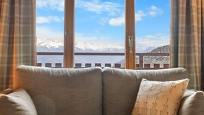 Een bank met een kussen in een kamer met uitzicht op besneeuwde bergen, te zien door een raam met gordijnen.