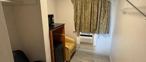 A small, narrow room with a tiled floor, a black office chair, a desk with a computer, a window with curtains, and a clothes drying rack.