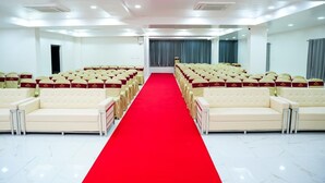 Une salle de conférence avec des rangées de canap és blancs et une moquette rouge, des unités de climatisation et une grande fenêtre avec des rideaux.