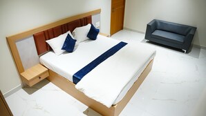 Un lit bien fait, avec une tête de lit en bois, une table de chevet et un canapé gris, dans une pièce moderne au sol recouvert de marbre blanc.