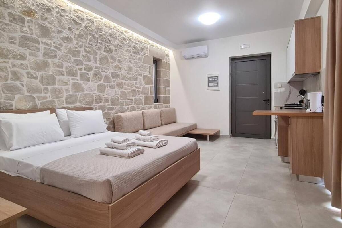 A bedroom with a stone wall, a bed with white linens, a wooden bench, and a kitchenette area.