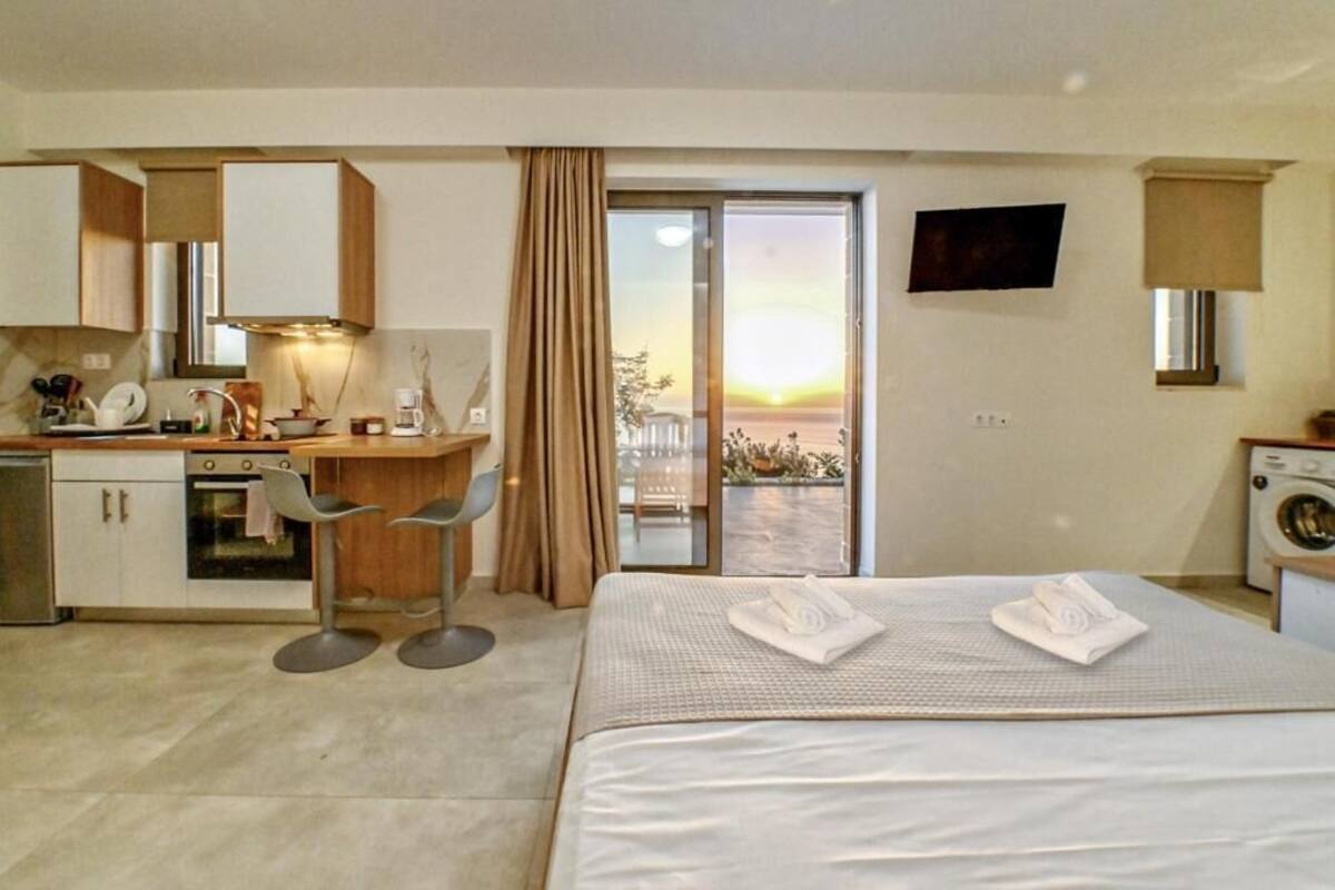 A hotel room with a bed, a desk, a chair, a washing machine, and a view of the sunset through the window.
