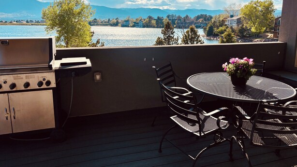 A balcony with a grill, a round table with chairs, and a view of a lake and mountains. - Spacious 4-bedroom ranch-style home 10 min from USAFA with lake & mountain views (Monument)