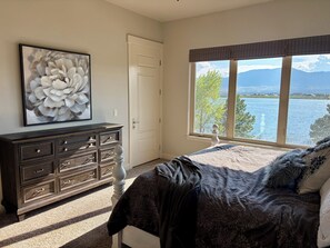 Ein Schlafzimmer mit Bett, Schminktisch, einem großen Fenster mit Blick auf einen See und Berge sowie einem Bild an der Wand.