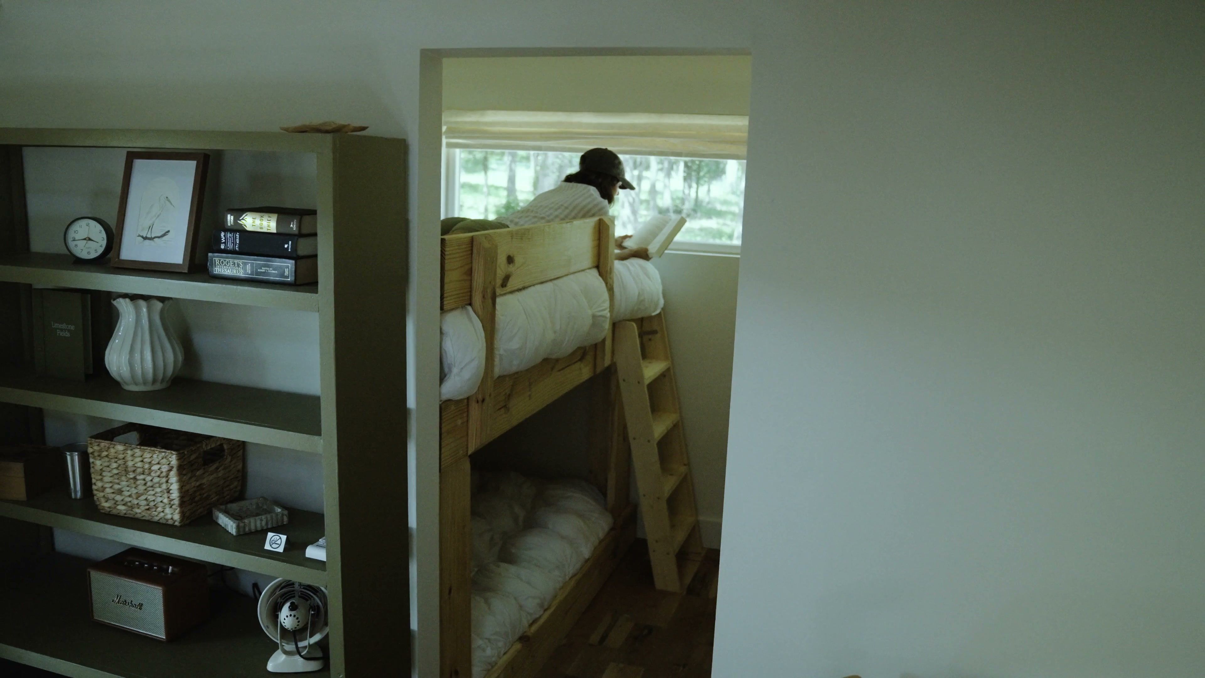 A room with a bunk bed, a bookshelf with various items, and a window with a view of trees.