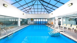 Une piscine intérieure dotée d’un toit en verre, de chaises transparentes placées au bord du bassin et offrant une vue sur la mer.
