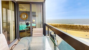 Un balcon avec une table en verre, une vue sur la plage et une chambre avec un lit et un miroir.
