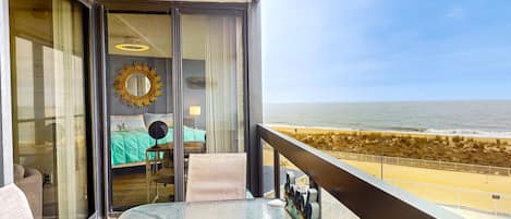 A balcony with a glass table, a view of the beach, and a room with a bed and a mirror.