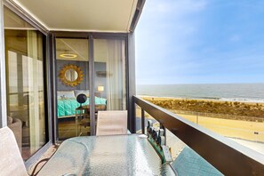 Ein Balkon mit Glastisch, Blick auf den Strand und ein Zimmer mit Bett und Spiegel.