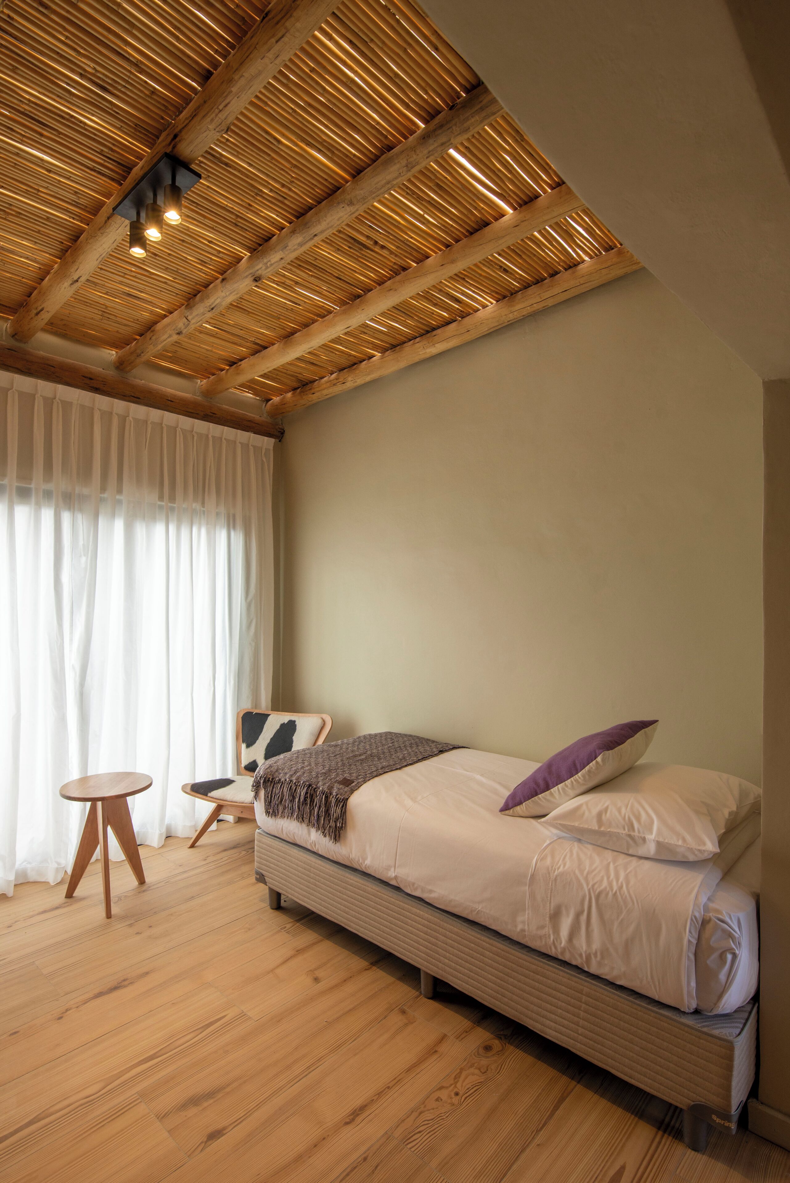 Een slaapkamer met een houten plafond, een bed met kussens, een stoel en een klein tafeltje.