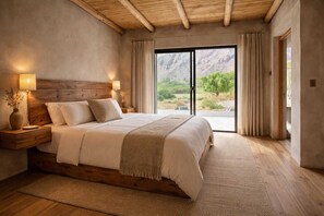 A bedroom with a large bed, wooden headboard, bedside lamps, and a view of a mountainous landscape through a sliding door. - Alma de Purmamarca (Purmamarca)