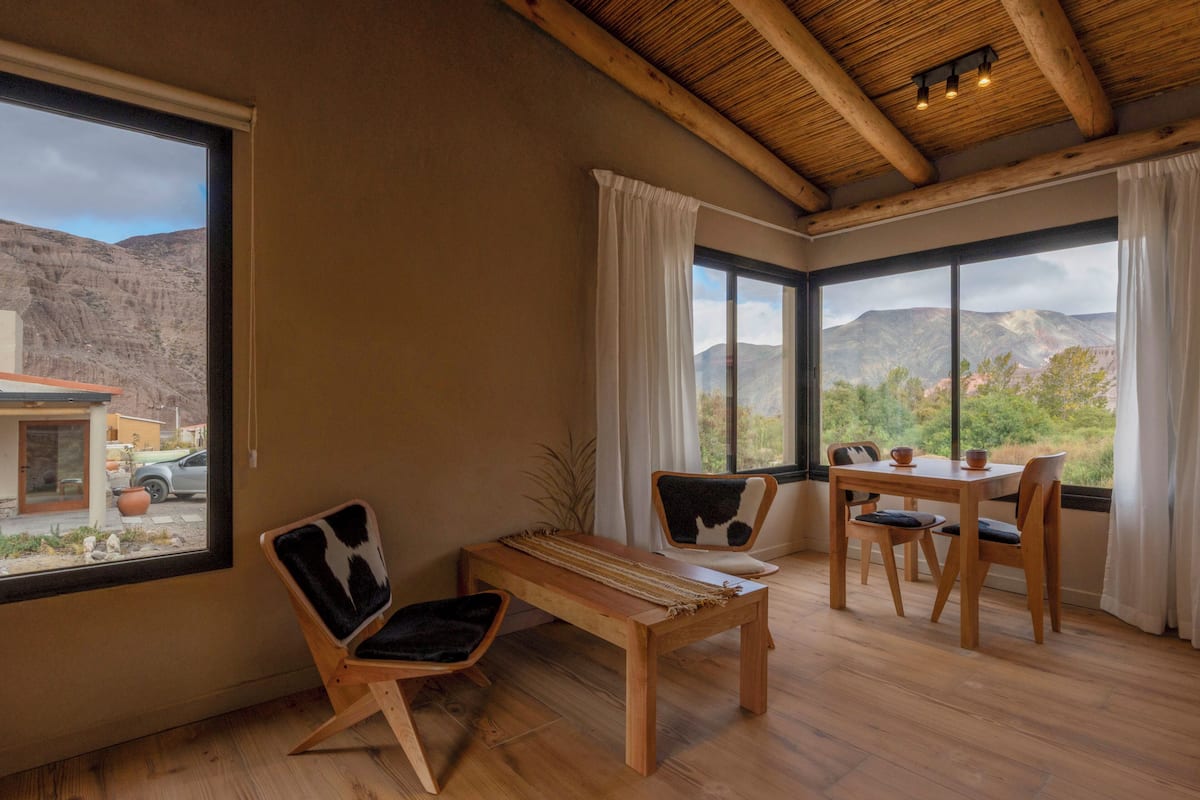 Una habitación con un banco de madera, dos sillas, una mesa y ventanas amplias con vistas a la montaña.