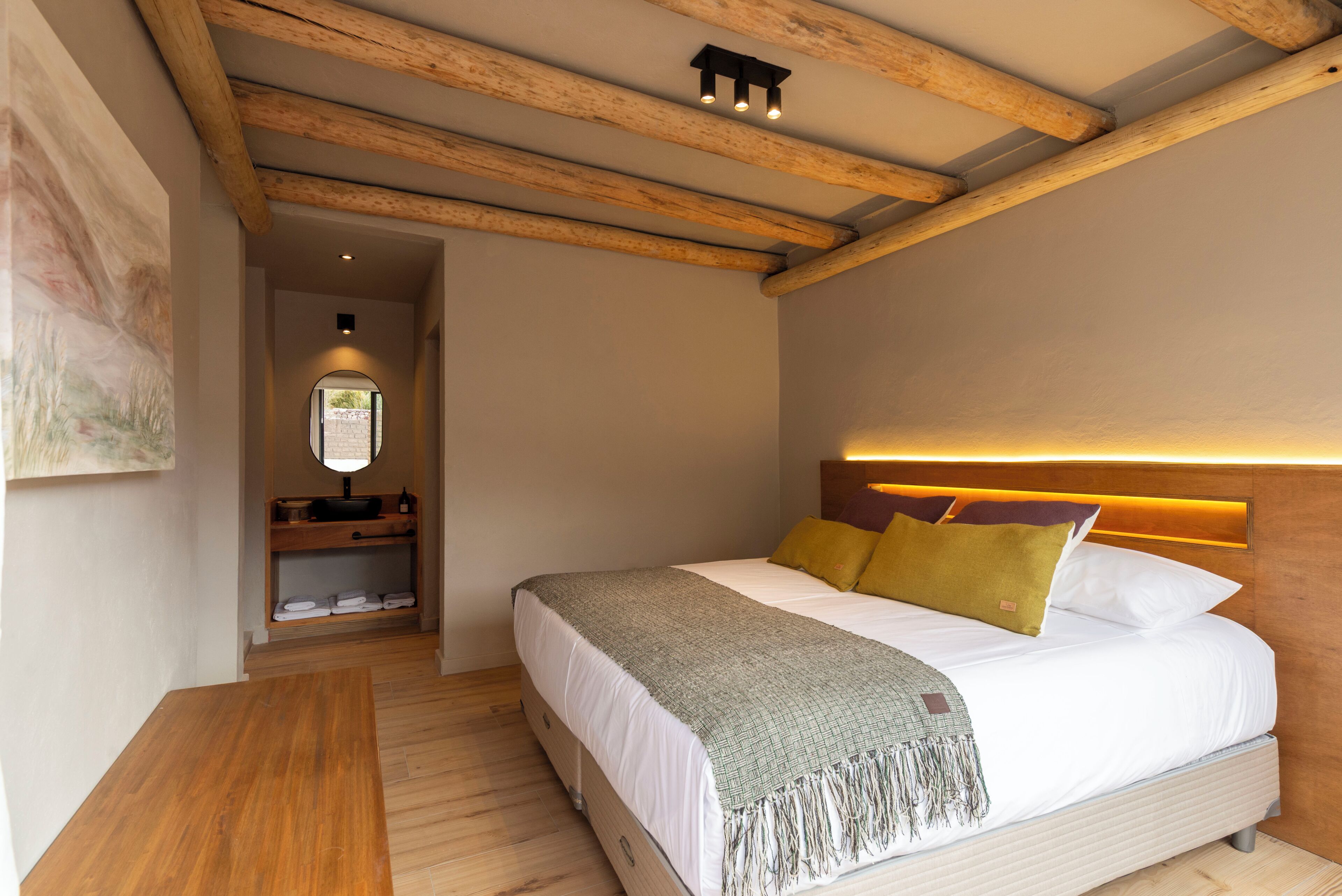 A bedroom with a wooden ceiling, a bed with white bedding and a green throw, a bedside table, and a mirror.