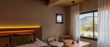 Un dormitorio con cama, una mesa de madera, dos sillas, una ventana con vistas a la montaña y un cuadro en la pared.