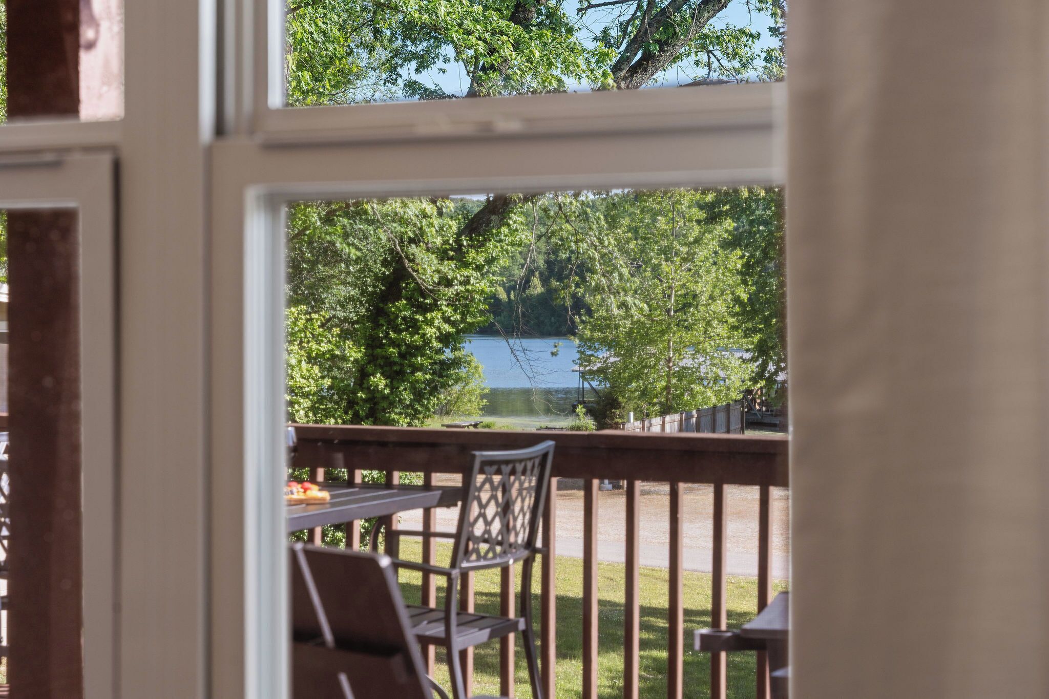 A view from a room with a wooden deck, a table, and a chair, overlooking a lake and trees.