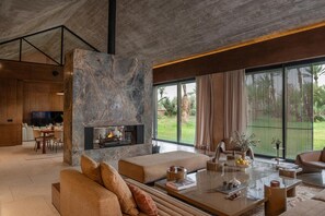 Ein modernes Wohnzimmer mit Kamin aus Naturstein, gemütlicher Sitzecke, Essbereich und großen Fenstern mit Blick auf Grünflächen.