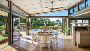 A modern dining area with a round table, white chairs, and a view of a swimming pool and greenery. - Glenbrook House and Studio (Ewingsdale)