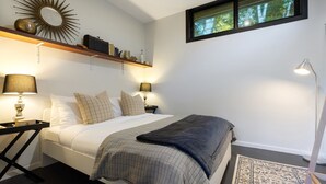 A neatly arranged bedroom with a bed, bedside tables, a nightstand, a lamp, a shelf with books and decorative items, a mirror, and a window with a view of trees. - Glenbrook Guest House (Ewingsdale)