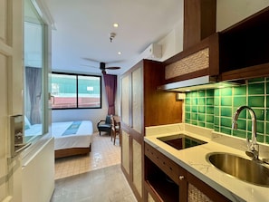 Quarto de hotel com cama, escrivaninha, cadeira, janela com vista, ventoinha no teto e área de cozinha com pia e bancada.