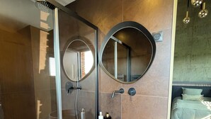 A modern bathroom with a glass shower enclosure, a round sink with a wooden bowl, two wall-mounted mirrors, and a bed with a green bedspread in the adjacent room. - La Gazolina The essence of calm, the luxury of the moment. (Boëseghem)