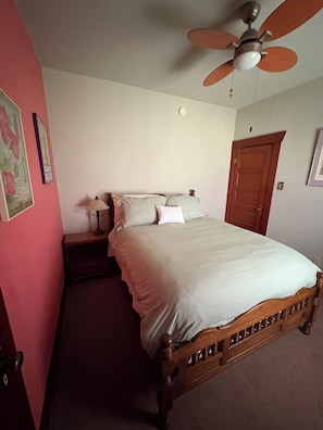 Une chambre à coucher avec un lit, une table de chevet, un ventilateur de plafond, une porte et un mur orné d’un tableau.