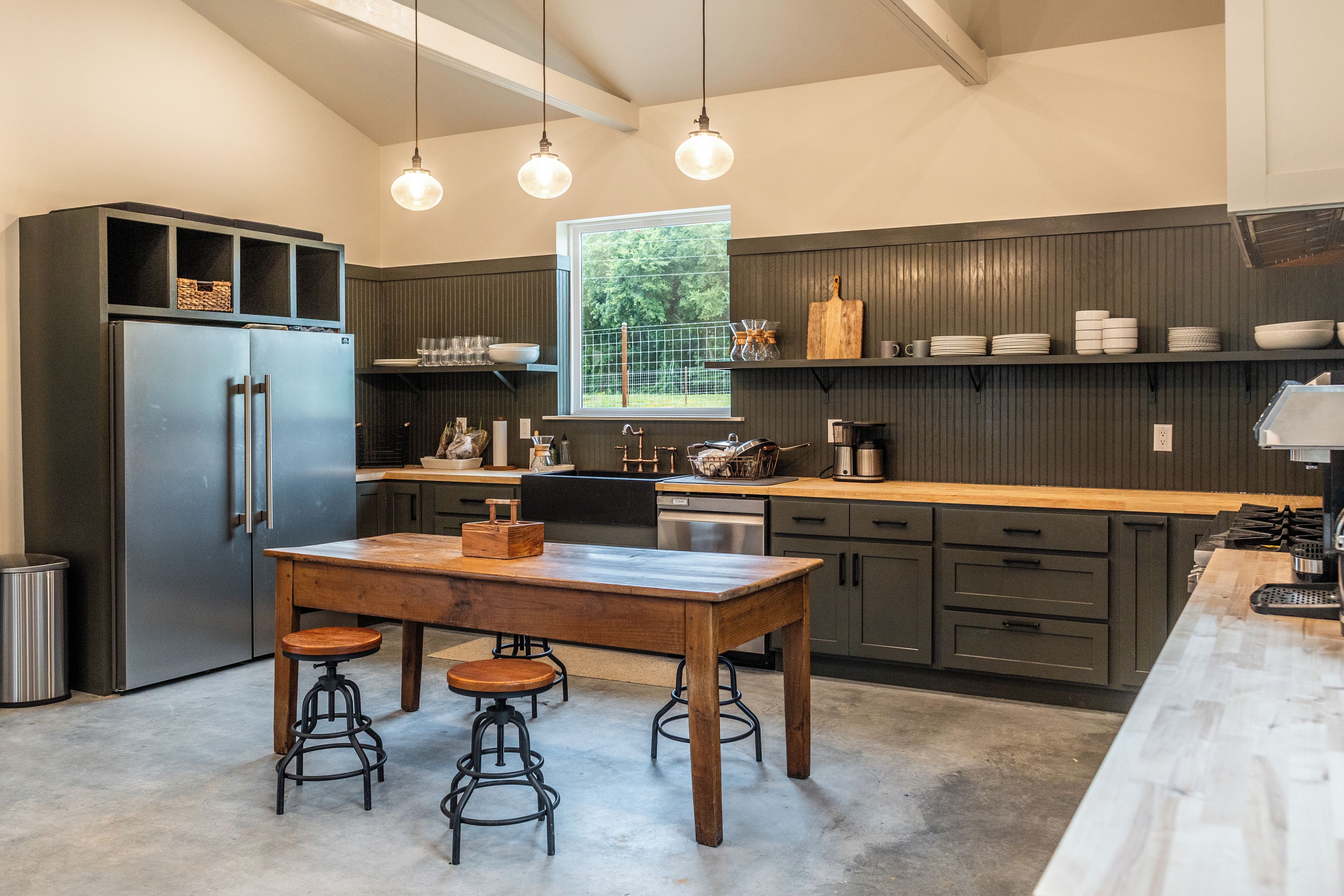 A modern kitchen with a wooden table, bar stools, a refrigerator, and a window with a view of greenery.