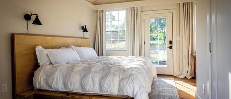 Une chambre à coucher avec un cadre de lit en bois, du linge de lit blanc, une lampe fixée au mur et une vue à travers une porte vitrée.