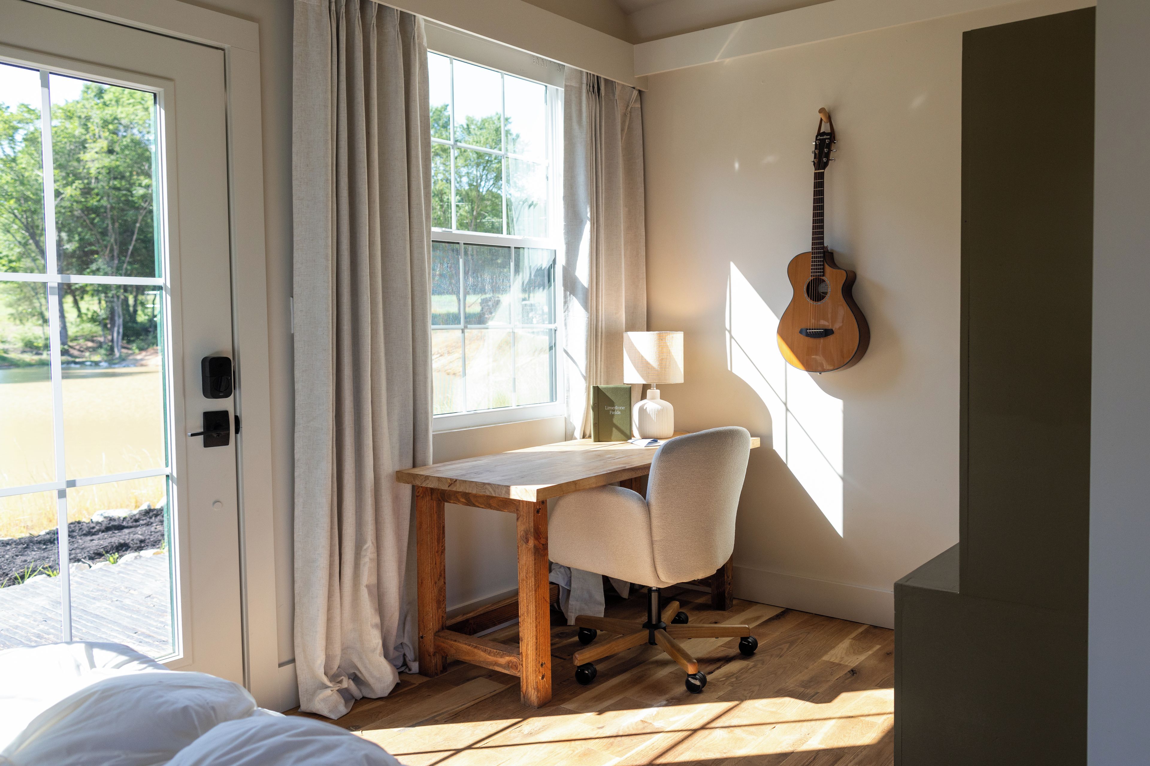 A room with a wooden desk, a chair, a guitar, and a window with curtains.