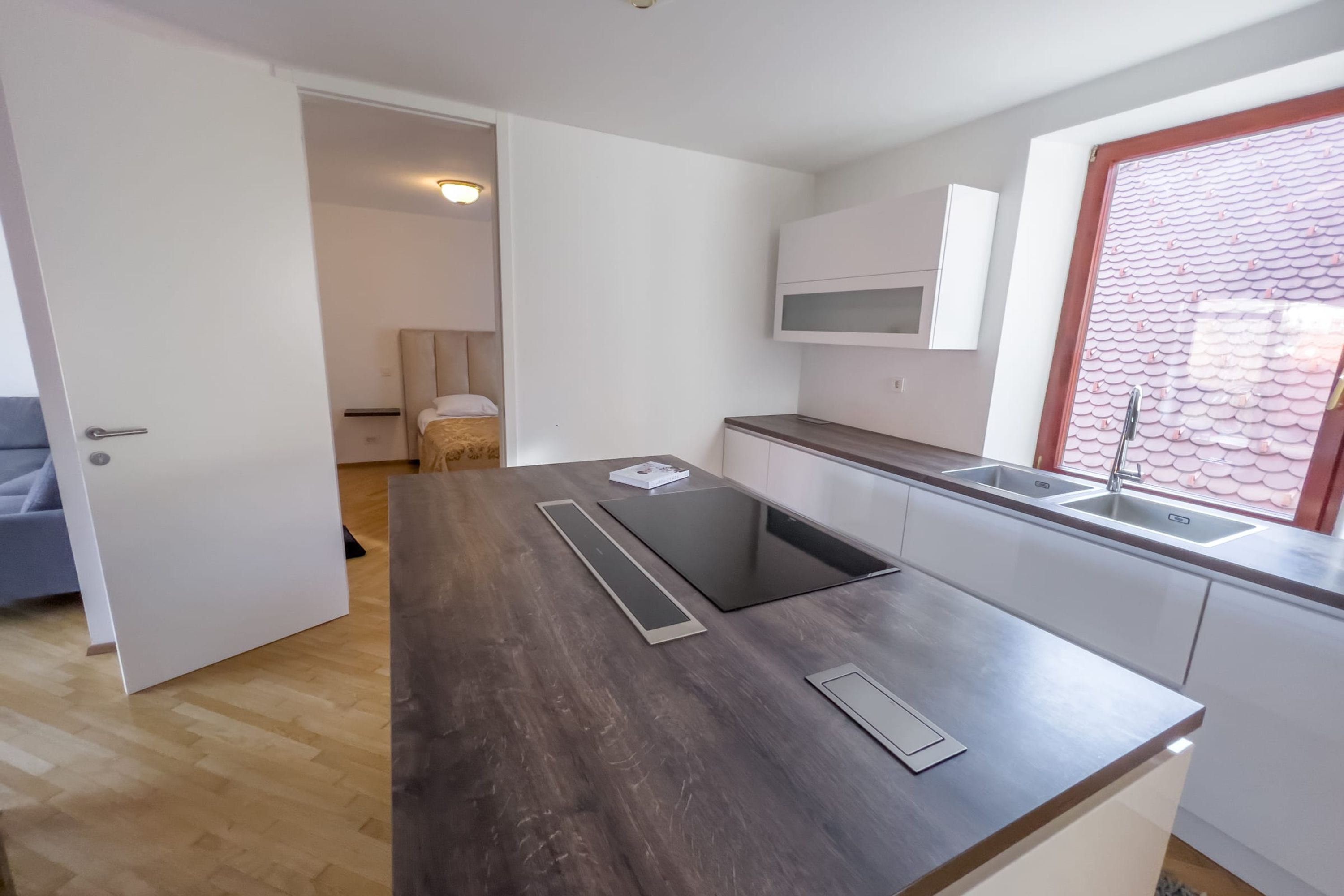 Cocina moderna con encimera oscura, armarios blancos y ventana con vistas a tejas rojas.