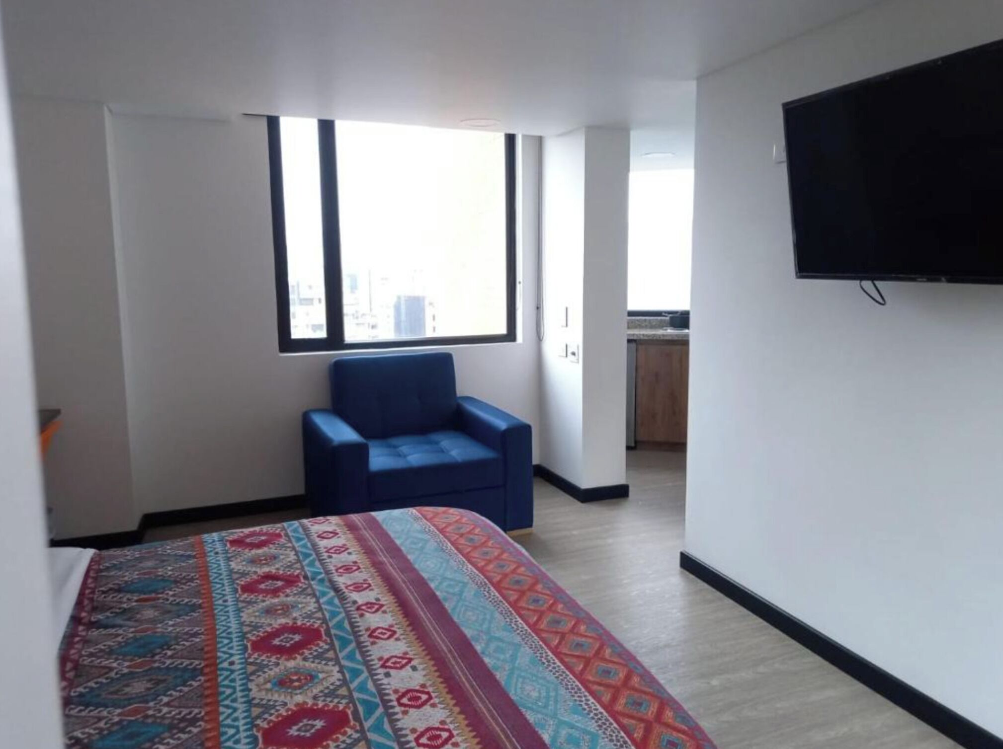 A hotel room with a bed, a blue chair, a TV, a window, and a view of the city.