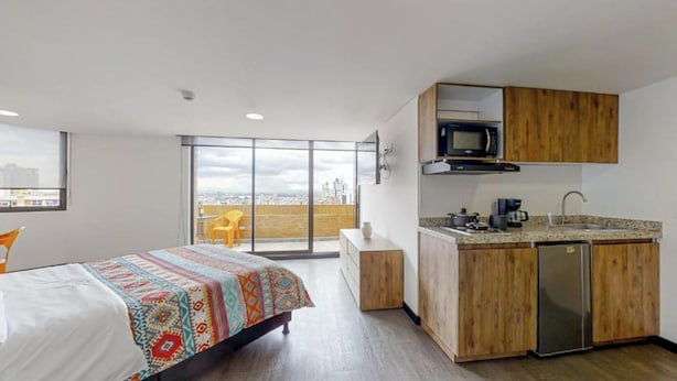 Una camera d'albergo moderna con un letto, un angolo cottura, una porta scorrevole in vetro su un balcone e vista sulla città.