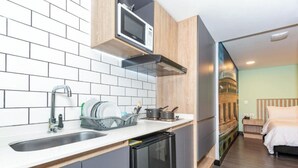 Angolo cottura compatto con forno a microonde, lavandino e scolapiatti. Si nota un piccolo frigorifero, e l'ambiente si apre su una camera da letto con un letto e un comodino.