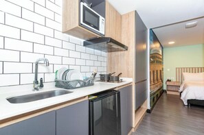 A compact kitchen with a microwave, sink, and dish drying rack. A small refrigerator is visible, and the room leads to a bedroom with a bed and a nightstand. - Terrazas (Bogota)