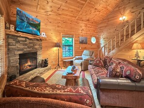 A cozy cabin interior with a stone fireplace, wooden walls, a comfortable sofa, and a television mounted above the fireplace. - A Bear's Eye View - Pet Friendly | Mountain View | Hot Tub | Game Room (Blue Ridge)