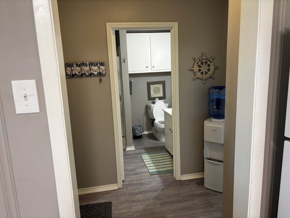 Un couloir étroit mène à une salle de bain équipée de toilettes, d’un distributeur d’eau et d’un tableau encadré accroché au mur.