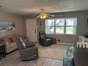Un salon comprenant un canapé, deux fauteuils, un ventilateur de plafond, une fenêtre donnant sur un beau paysage et une porte.
