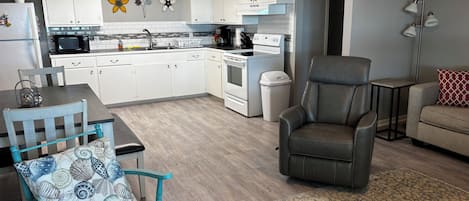 Une cuisine moderne avec des meubles blancs, un four à micro-ondes et une cuisinière. On y trouve une table à manger avec des chaises, un canapé et un tapis à motifs.